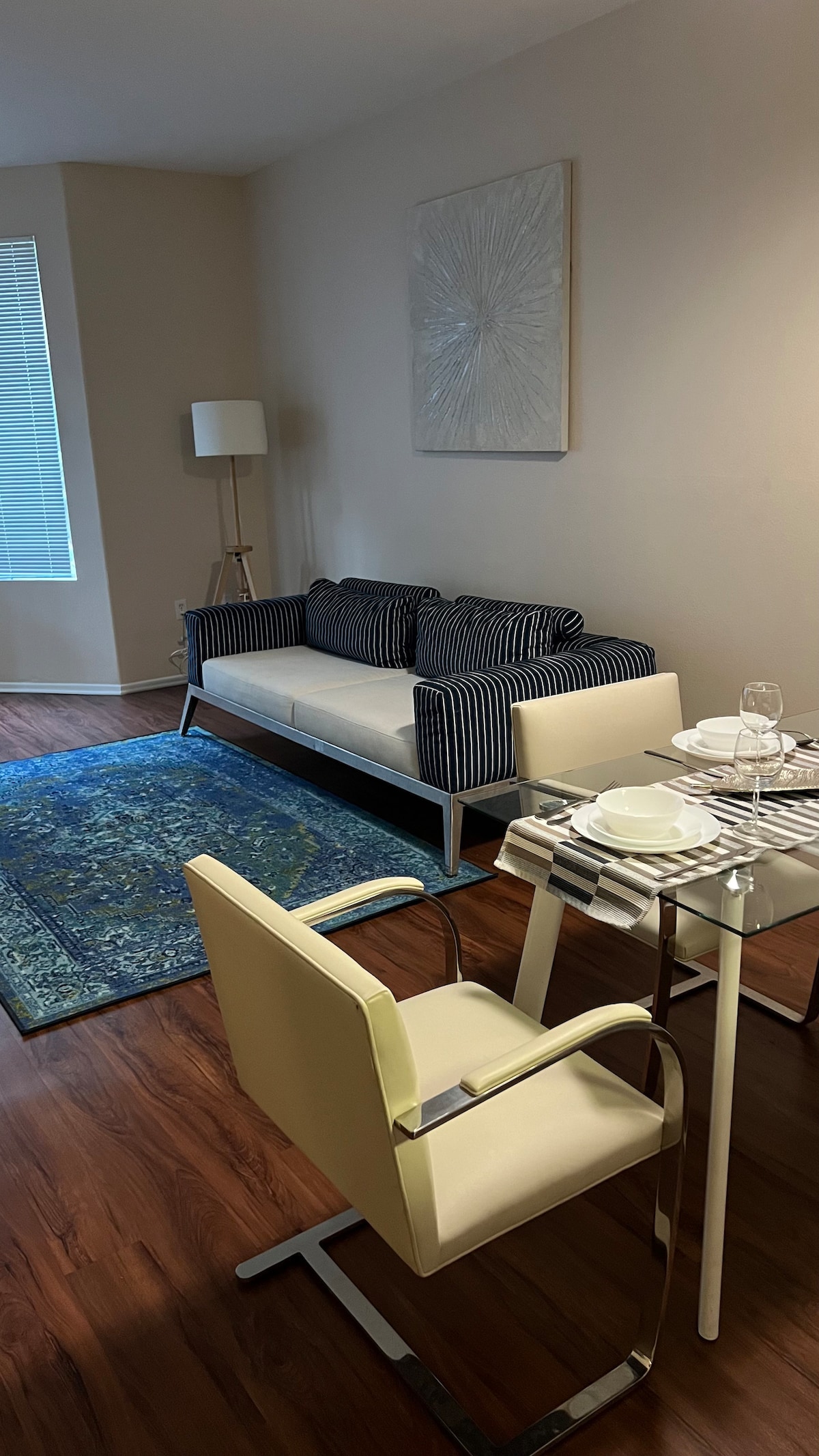 A well-lit living area showcases a modern sofa with striped cushions and a light-colored wall art piece. Nearby, a glass dining table is set with plates and glasses, complemented by two sleek, white chairs. A textured area rug adds a touch of warmth beneath.