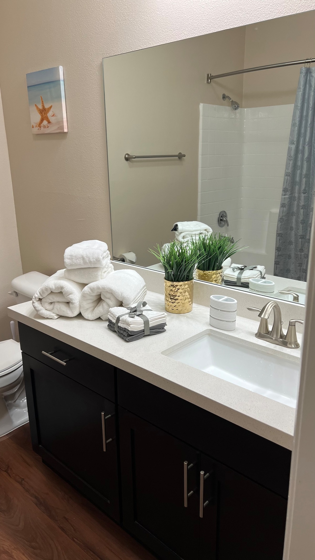 A well-appointed bathroom features a neatly arranged stack of white towels beside a small potted plant on a dark wooden countertop. A large mirror reflects the space, which includes a shower area and a modern faucet above the white sink.