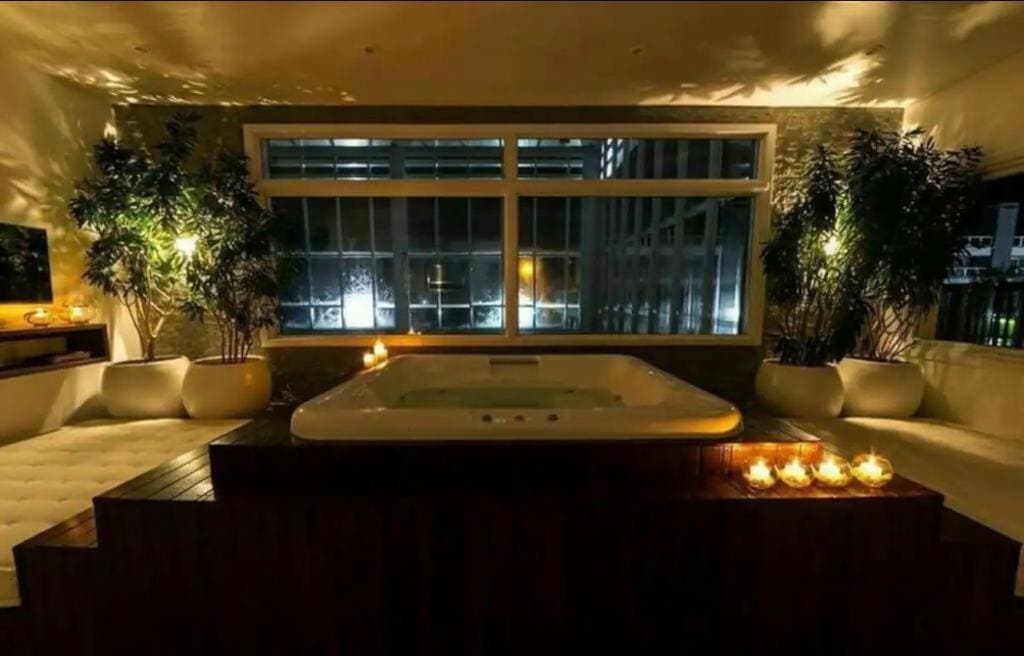 An indoor jacuzzi is surrounded by potted plants, illuminated by candlelight. A large window allows for natural light, showcasing a tranquil ambiance. Soft lighting highlights the wooden platform beneath the jacuzzi, creating a serene and comfortable environment.