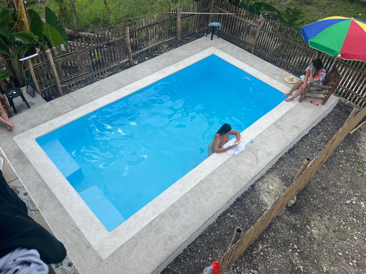 A rectangular swimming pool is surrounded by a smooth concrete deck. Guests are visible lounging on the side, with one person relaxing in the water. Colorful umbrellas provide shade, and a rustic bamboo fence encloses the area, creating a peaceful outdoor space.