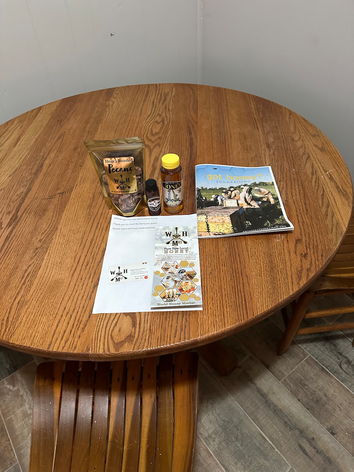A wooden round table is displayed, featuring a variety of items. A bag of coffee beans, two bottles of condiments, and a brochure are arranged on the surface, along with a magazine showcasing outdoor scenery. Two wooden chairs are positioned around the table.