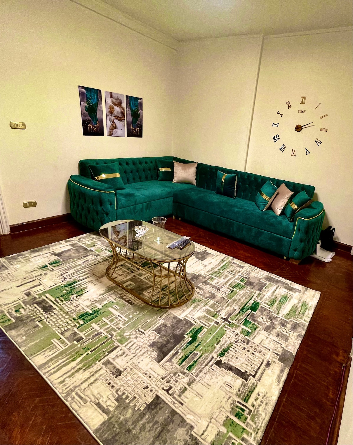 A spacious living room features a green sectional sofa with decorative cushions, arranged around a glass coffee table. A textured area rug adds warmth to the polished wooden floor. Three framed artworks are displayed on the wall, complemented by a stylish wall clock.
