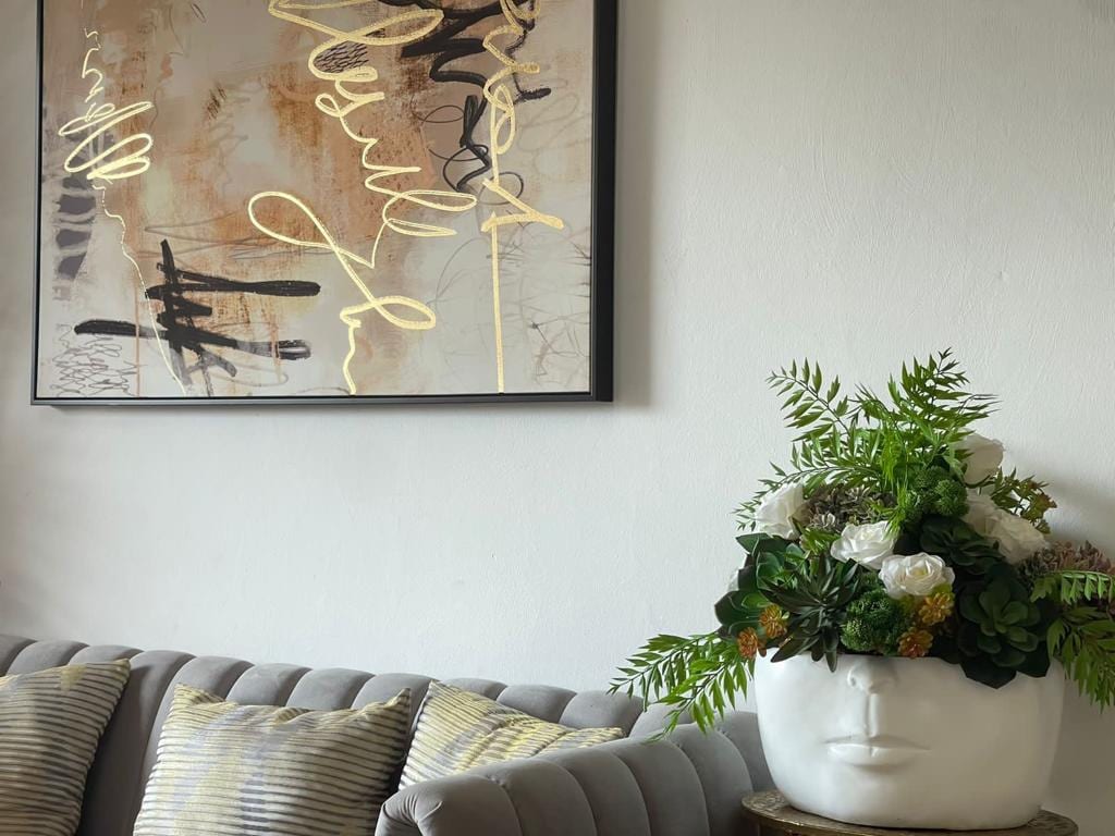 A modern living space features a plush gray sofa adorned with striped cushions. Above, an abstract art piece with gold accents is displayed on the wall. A unique planter in the shape of a face holds an assortment of greenery and white flowers, adding a decorative touch.
