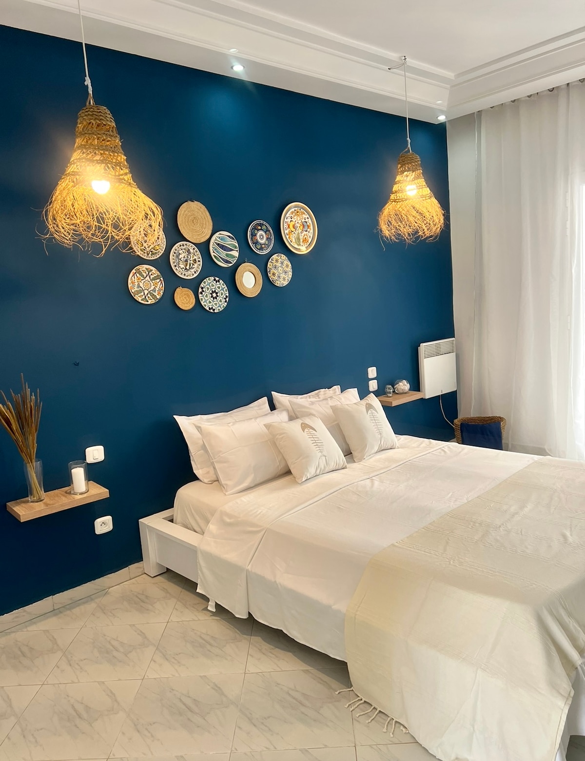 A calming bedroom features a deep blue accent wall adorned with decorative plates. Soft white bedding is layered over a spacious bed, with two pendant lights hanging overhead. Natural light enters through a sheer curtain, illuminating a minimalist bedside table.