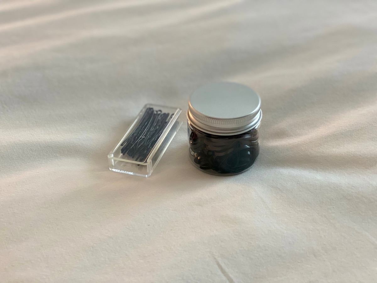 A transparent container holds a collection of hair ties, while a small box beside it contains assorted bobby pins. Both items are placed on a neatly made bed featuring smooth, light-colored bedding.