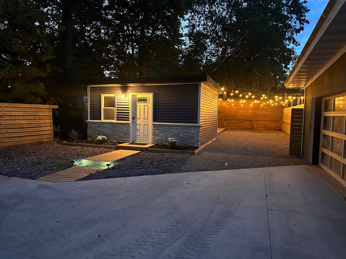 An inviting exterior is highlighted by soft lighting from string lights above, illuminating the pathway leading to a welcoming front door. The surrounding gravel area features neatly arranged plants, while a modern structure stands amidst a tranquil setting, with additional outdoor space visible in the background.