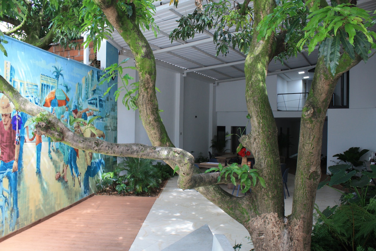 A vibrant mural created by local artists is displayed against a backdrop of greenery. A large tree branches out towards the viewer, complementing the outdoor area that features modern furnishings and a natural, tranquil ambiance.