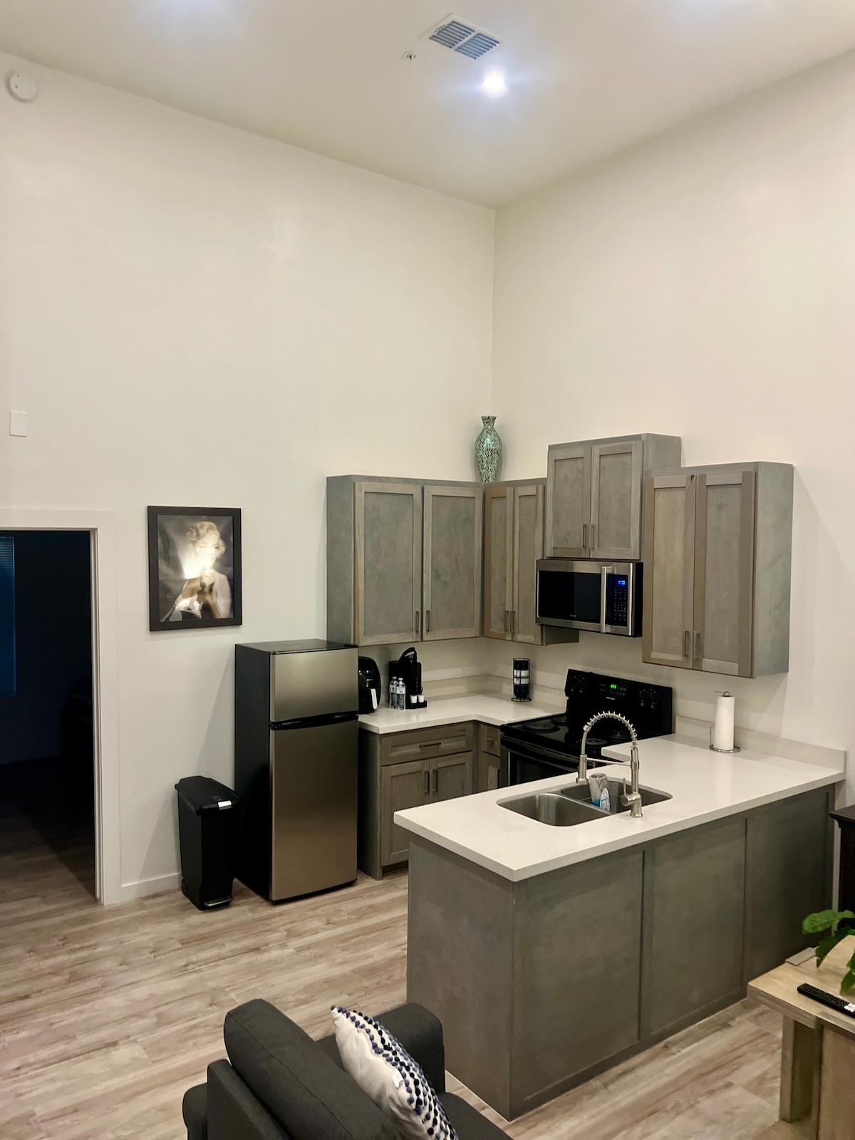 A modern kitchen is showcased with gray cabinetry and stainless steel appliances, including a refrigerator and oven. A countertop with a sink provides a functional space for meal preparation. Natural light brightens the area, which is accentuated by minimalistic decor and a cozy seating arrangement.