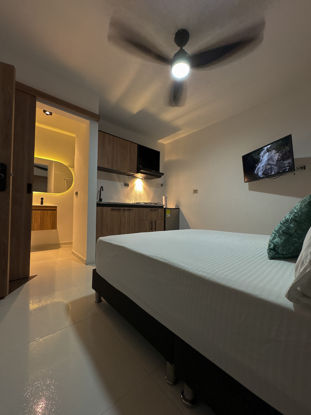 A modern studio space is showcased, featuring a spacious bed with crisp white linens and a decorative pillow. A kitchenette is visible in the background, equipped with wooden cabinetry and a compact dining area. Soft lighting creates a cozy ambiance, complemented by a wall-mounted television.