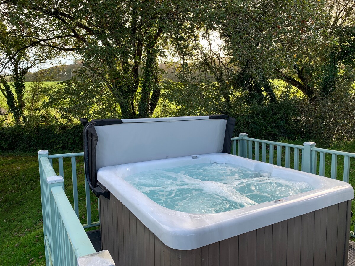 A private hot tub is set on a wooden deck, surrounded by lush greenery. The tub is filled with bubbling water, and the protective cover is positioned beside it. Sunlight filters through the trees, creating a peaceful outdoor setting.