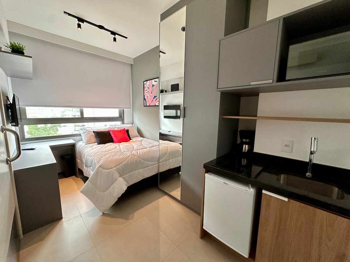 A modern studio is presented with a spacious double bed dressed in white linens, against a gray wall. A compact kitchenette features a dark countertop, a coffee maker, and a small refrigerator. Large windows provide natural light and a view of the outside.