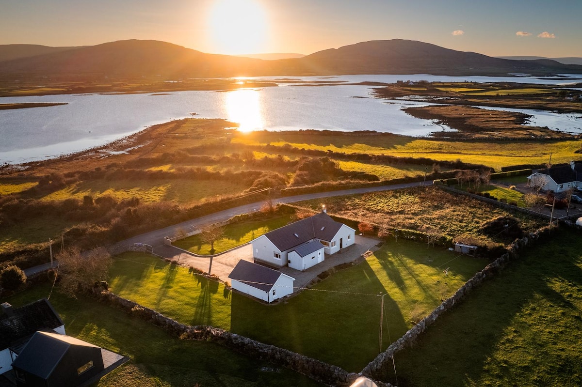 An aerial view captures the luxury cottage surrounded by lush green fields and scenic water. The sun sets in the background, casting a warm glow over the tranquil landscape. The property is framed by natural beauty, illustrating its serene location.