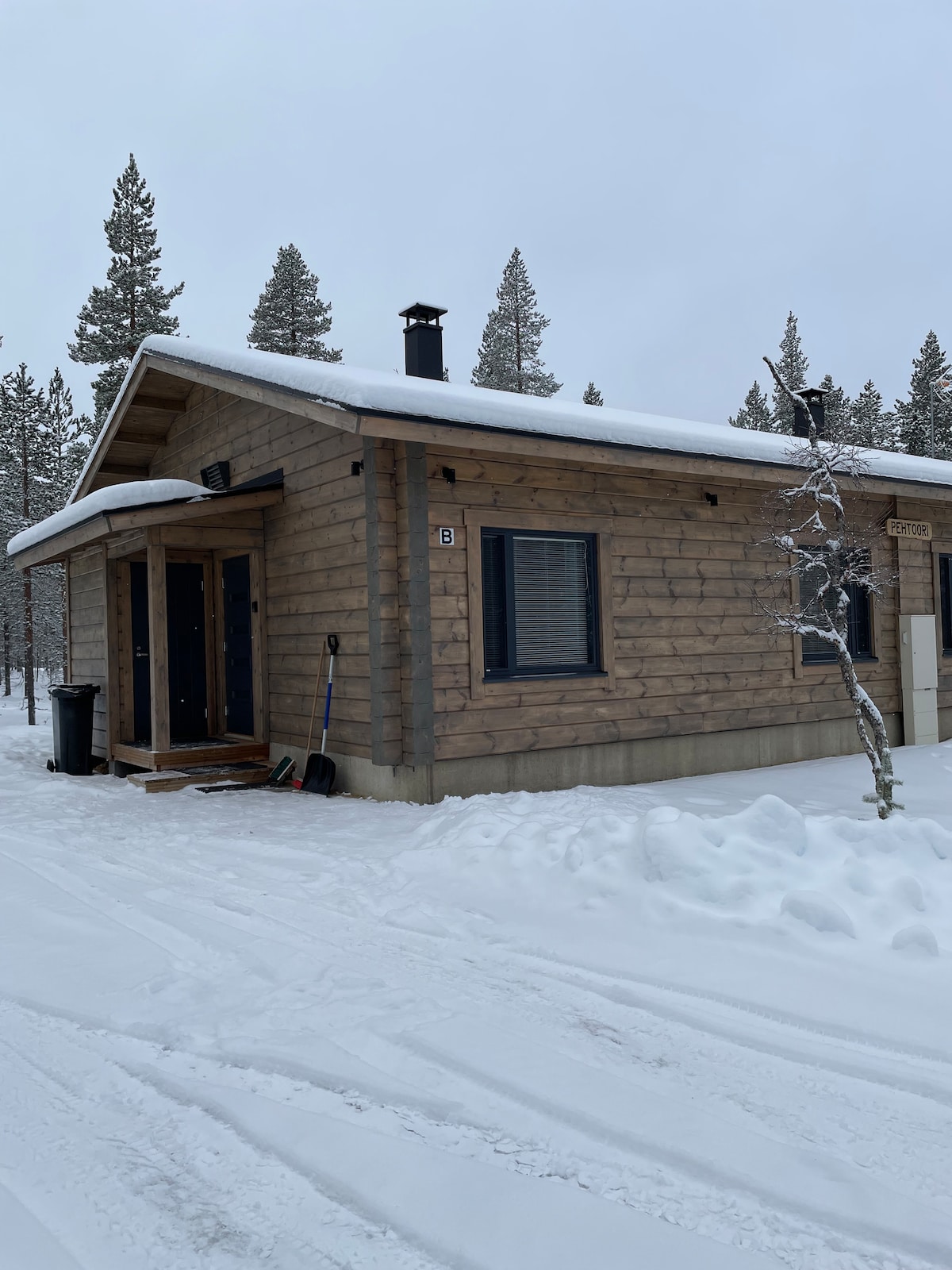A charming wooden cabin is situated among snow-covered trees, with a snow-dusted roof and a clear pathway leading to the entrance. The structure features dark window frames and a small porch, contributing to its rustic appeal in a serene winter setting.