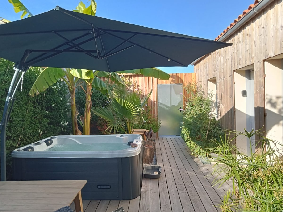 A private outdoor space features a hot tub under a canopy, surrounded by lush greenery and tropical plants. Wooden decking provides a natural ambiance, while a textured wall in the background contributes to the serene setting.