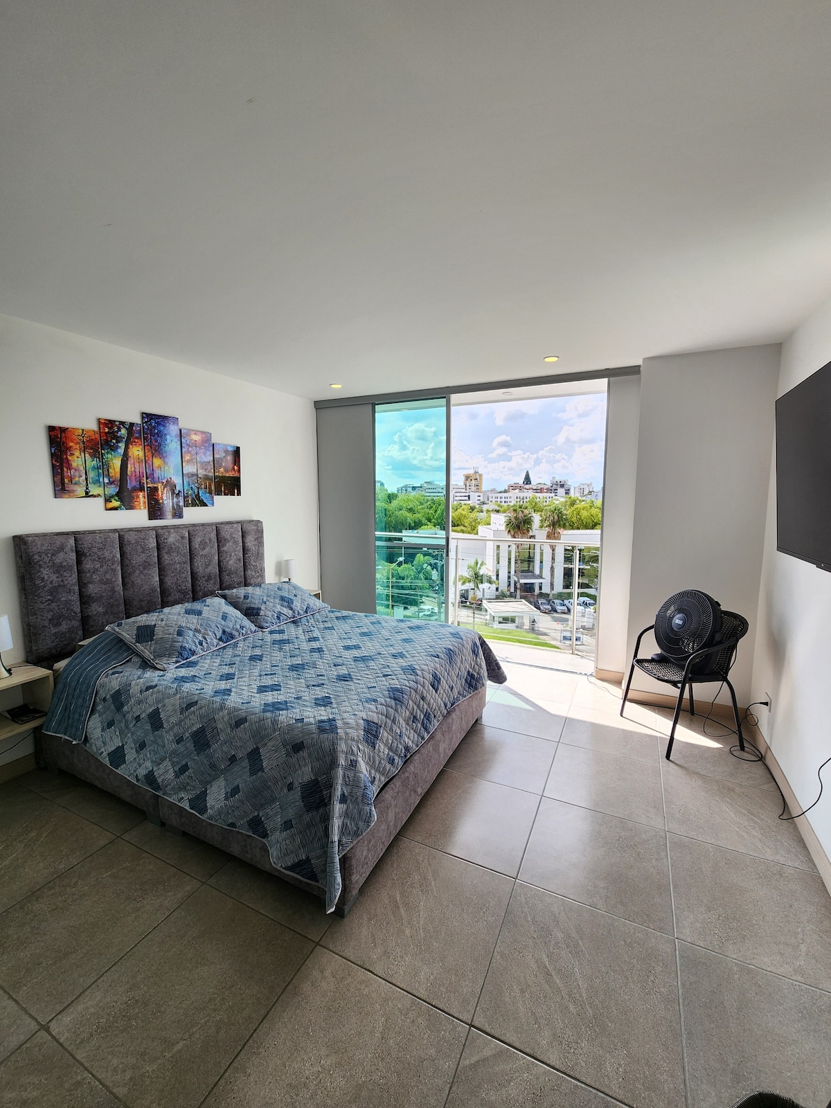 A modern bedroom features a spacious bed with a patterned blue blanket and decorative wall art. Large windows allow natural light to fill the room, providing a view of the outside landscape. A flat-screen TV is mounted on the wall, and a fan is positioned nearby.