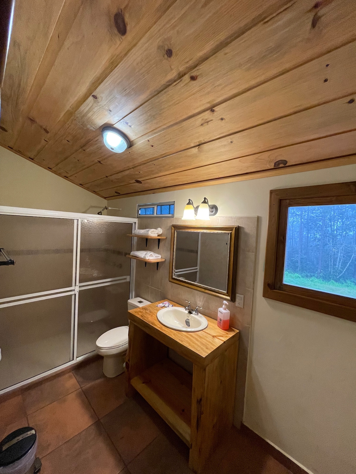 The bathroom features natural wood accents on the ceiling and a wooden vanity with a sink. A shower stall is enclosed with glass doors, and a mirror reflects the soft lighting. A small window allows natural light to enter, providing a view of the surrounding greenery.