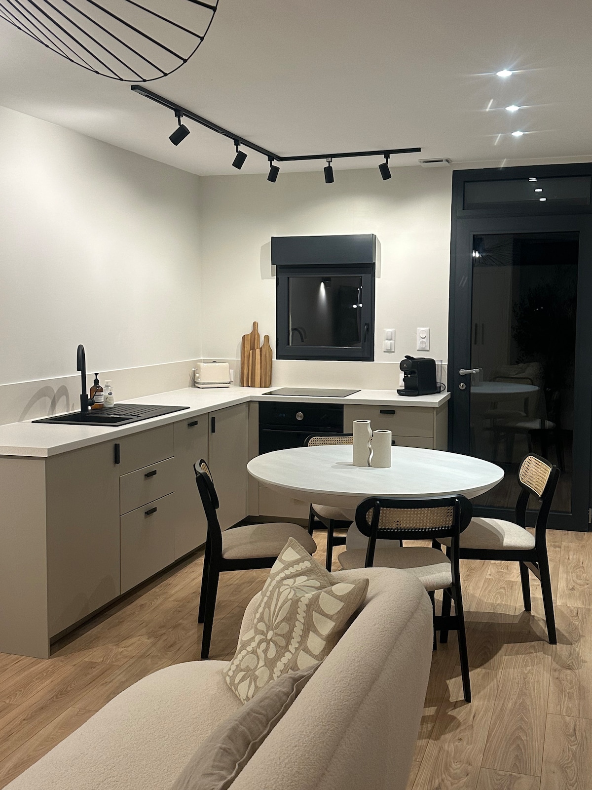 A modern kitchen area features sleek cabinets and a small round dining table surrounded by four chairs. A coffee maker is visible on the counter, complemented by light-colored decor. Large windows allow for natural light, and track lighting highlights the space.