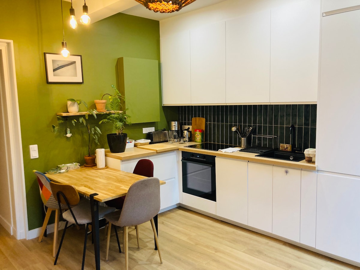 A modern kitchen area features a mix of green and white cabinetry, complemented by a wooden dining table surrounded by four chairs. A variety of plants adorn the shelves, and appliances include a stovetop, oven, and refrigerator.