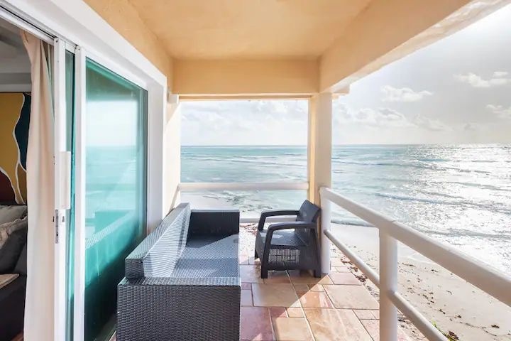 The balcony area features a comfortable seating arrangement with two chairs, positioned to offer direct ocean views. Natural light floods the space, enhancing the connection to the serene beach below. The ocean waves are visible, creating a calming atmosphere.
