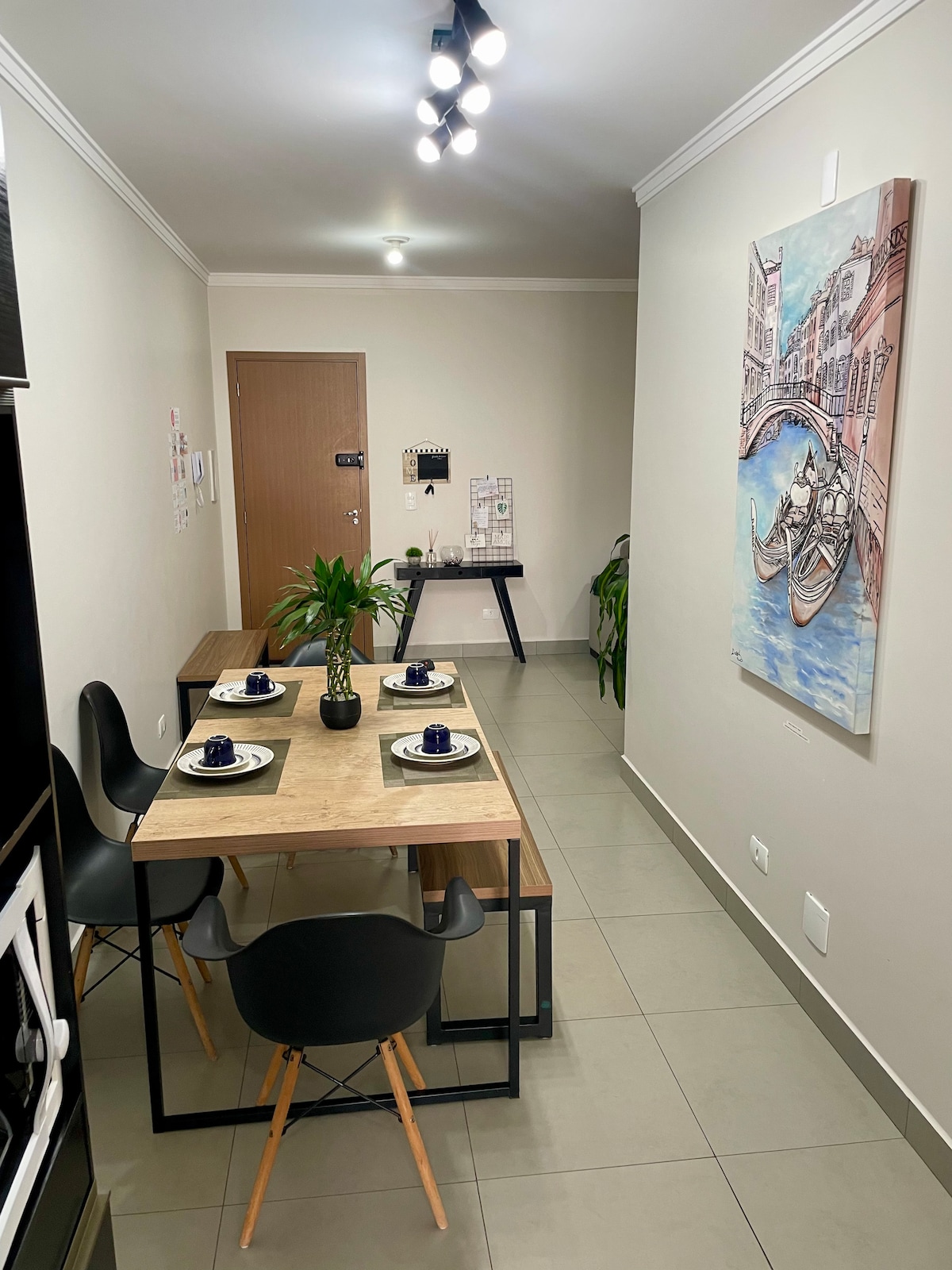 A dining area features a wooden table surrounded by four black chairs. A decorative plant sits at the center of the table, accompanied by place settings with blue plates and white napkins. A wall art piece depicting a canal scene adds interest to the light-colored walls.