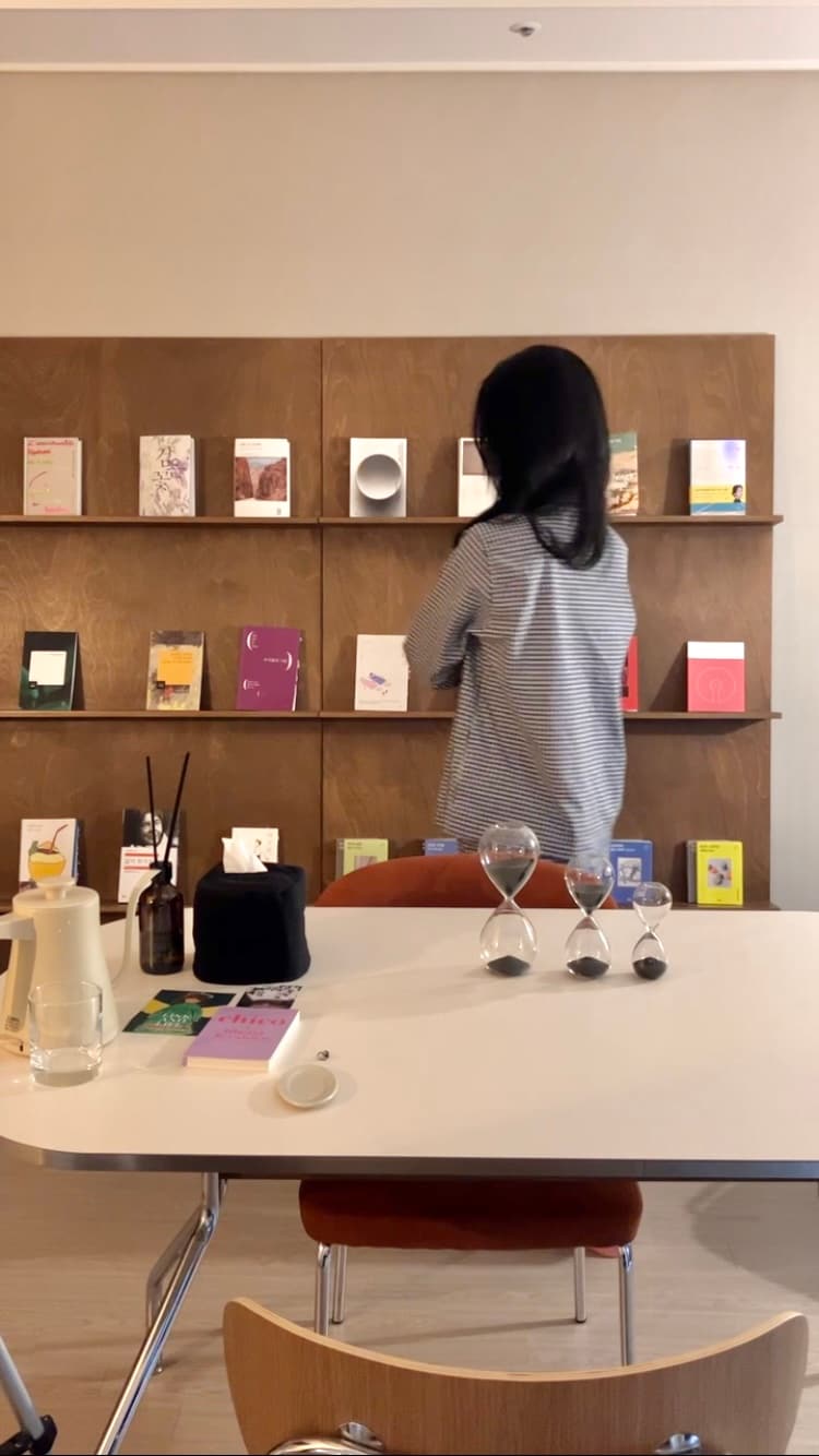 A wooden wall shelf displays an array of books and art pieces in various colors and designs. A white table features a few decorative items, including hourglasses and a neutral-colored pitcher. A person stands in a striped shirt, examining the shelf.