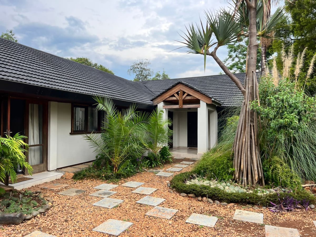 An entrance path lined with stone pavers leads to a charming home, framed by lush greenery and tropical plants. A wooden canopy accents the doorway, providing a welcoming focal point, while the surrounding landscape features a mix of pebbles and vibrant foliage.