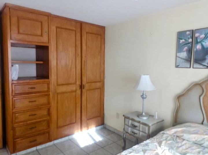 A cozy sleeping area is presented with light-colored walls and tiled flooring. A spacious wardrobe with wooden finishes provides ample storage alongside a decorative nightstand. A soft lamp offers ambient lighting, complementing the gentle tones of the bedding and wall art depicting floral designs.