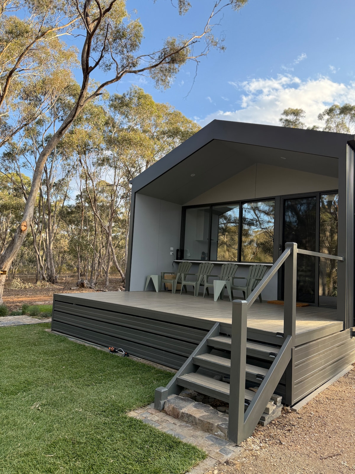 A modern cabin exterior is showcased with a deck leading up to the entrance. Large windows invite natural light, and a dining area with chairs can be seen inside. Surrounding bushland adds to the serene environment.
