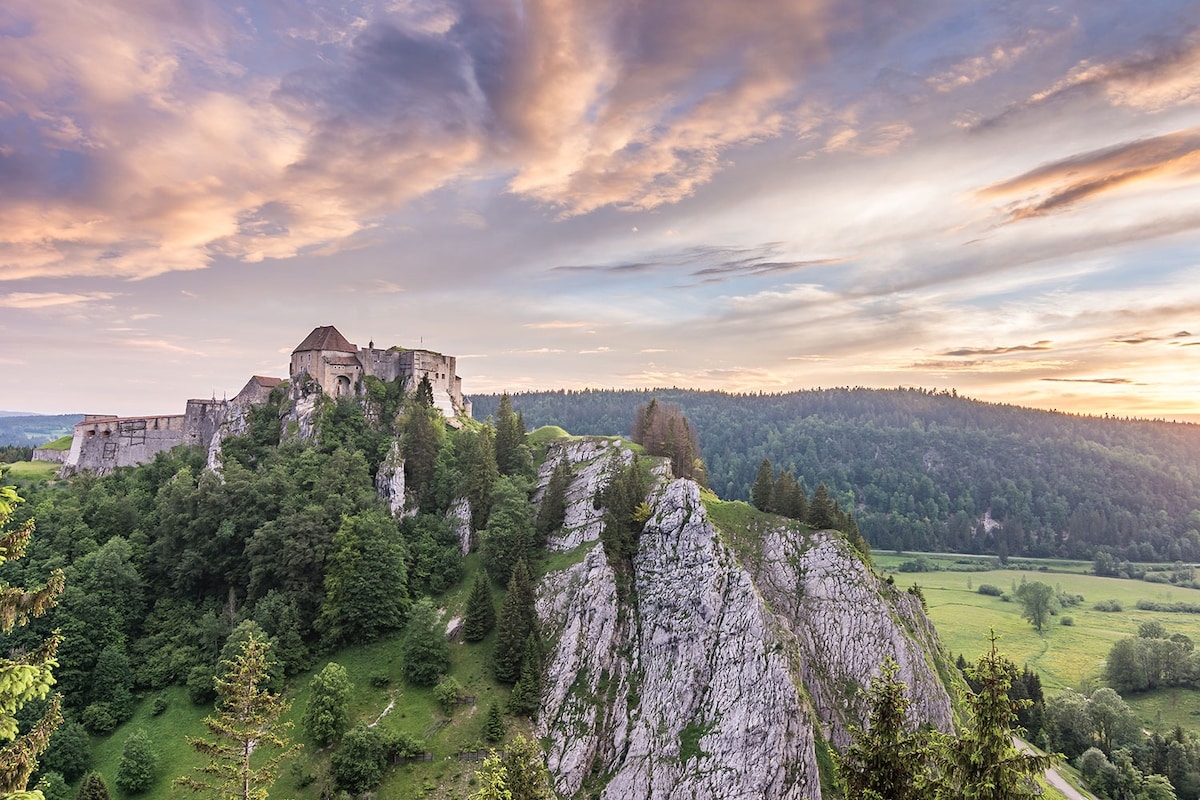 A historic castle is perched atop a rocky hill, surrounded by lush greenery. The landscape features rolling hills in the background, with a sky painted in soft shades of orange and purple as the sun sets.