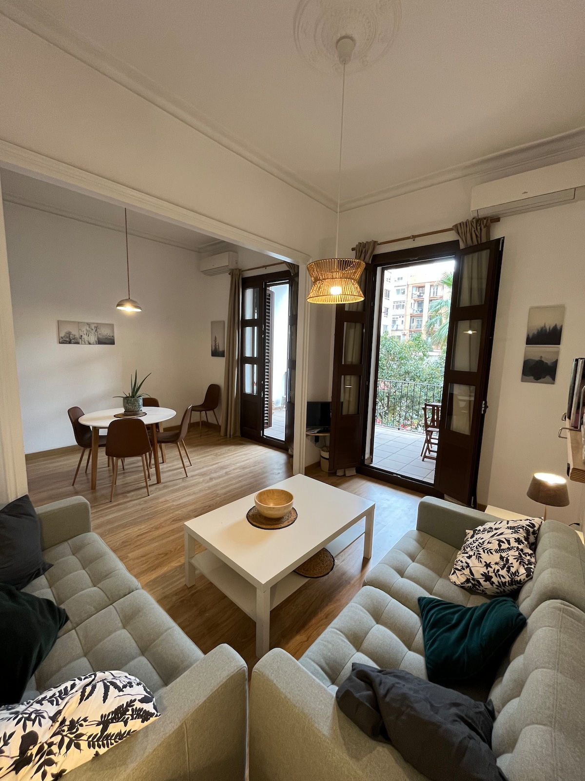 A bright and spacious living area features two comfortable sofas, a central coffee table, and a dining table surrounded by chairs. Natural light floods in through large windows, offering views of greenery outside. Decorative cushions add a touch of comfort to the space.