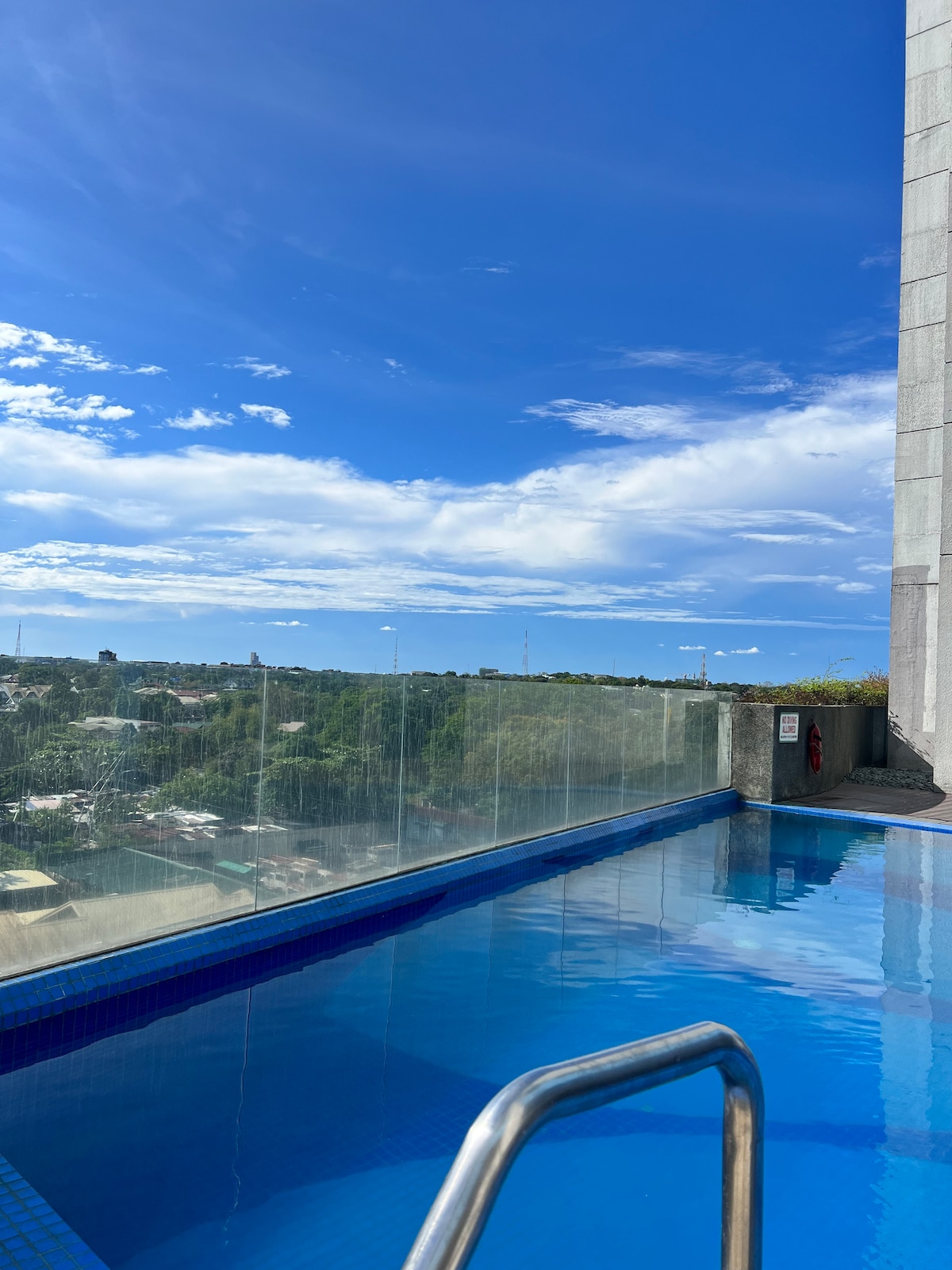 The rooftop pool features clear blue water reflecting the expansive sky. A glass barrier provides unobstructed views of the surrounding cityscape and greenery. Bright sunlight illuminates the scene, creating a serene setting for relaxation.