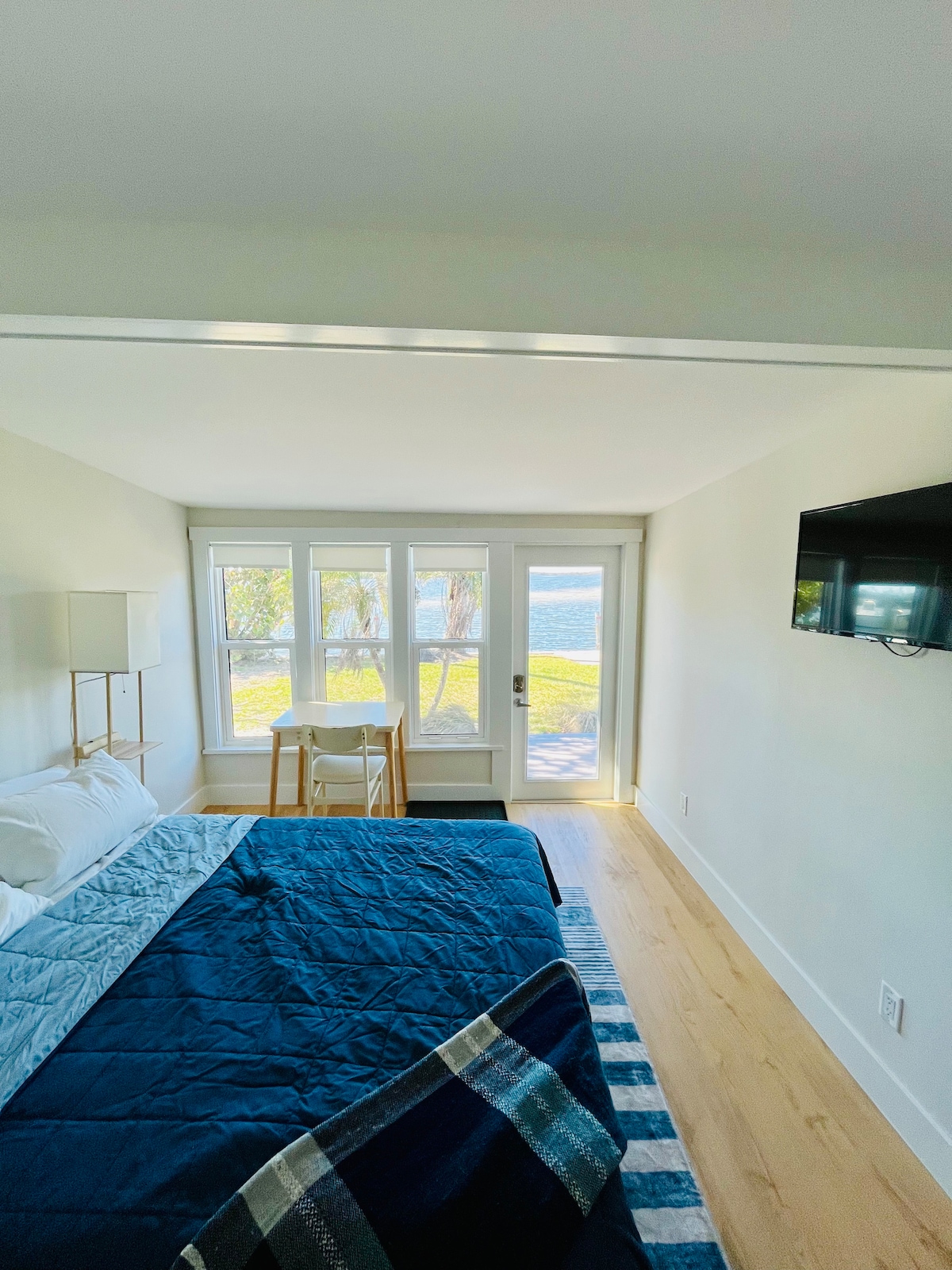 The serene bedroom features a spacious bed draped in a light blue coverlet. A set of large windows provides a view of the open waters, while a small dining nook is positioned nearby. A flat-screen TV is mounted on the wall opposite the bed.