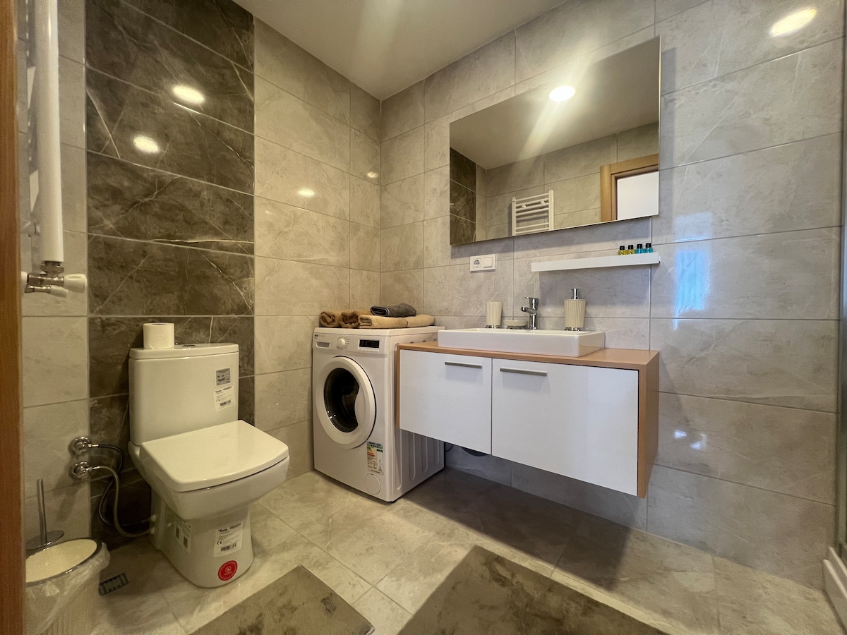 The bathroom features modern tiles in neutral tones. A sleek vanity with a sink is positioned against the wall, accompanied by a mirror above. A washing machine is located adjacent to the vanity. A towel rack and toiletries are visible, enhancing the functionality of the space.