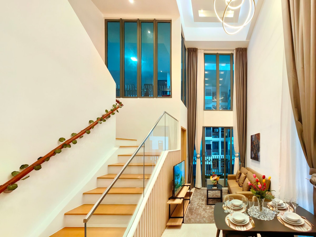A modern duplex loft showcases a spacious living area with high ceilings and large windows allowing natural light. A stylish staircase leads to the upper level, while a neatly arranged dining table is set nearby. Soft furnishings and decorative elements are present, contributing to a contemporary ambience.
