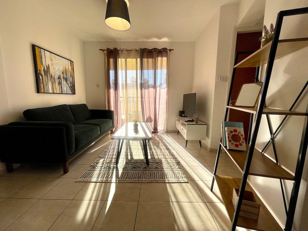A cozy living area features a dark green sofa paired with a patterned area rug. A coffee table sits at the center, flanked by a television and a light wood shelf displaying decor. Natural light streams through the floor-to-ceiling curtains, enhancing the space.