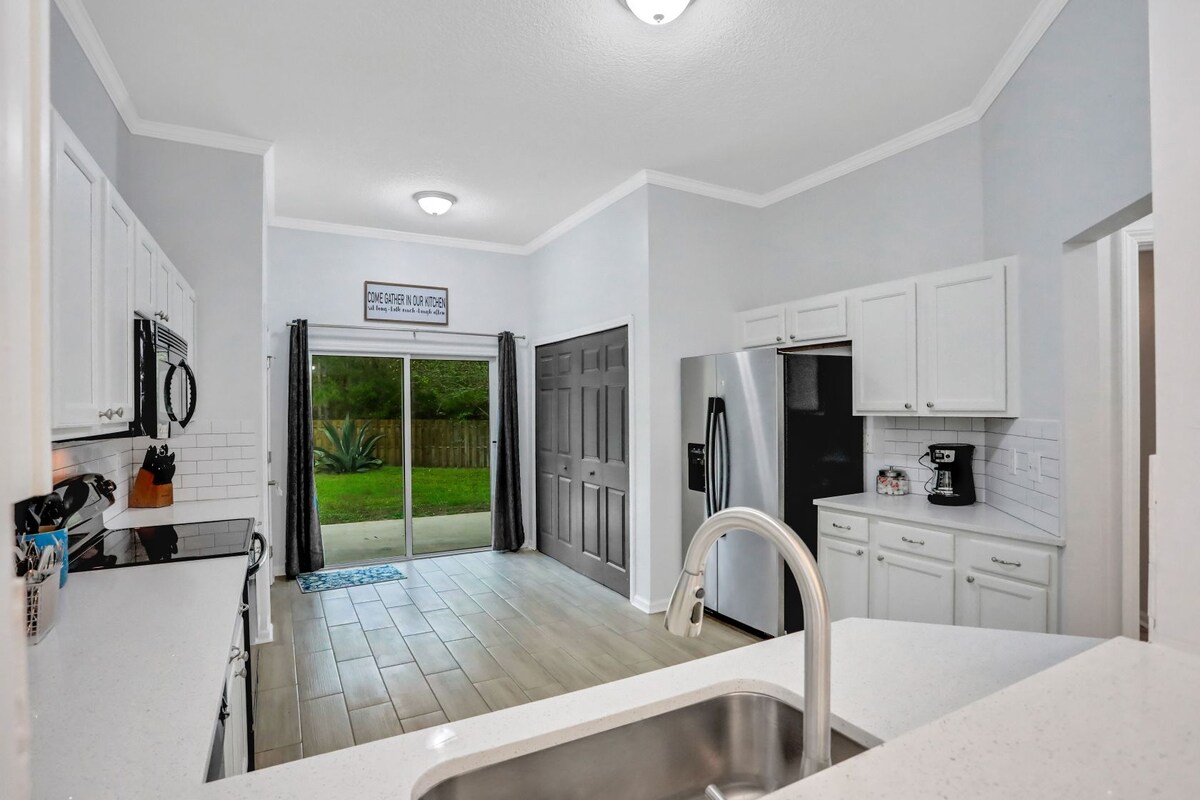 The kitchen features white cabinetry and a spacious countertop. A stainless steel refrigerator and coffee maker are visible. Natural light enters through sliding glass doors, leading to a serene outdoor space. The floor is finished with light-colored tiles for an airy feel.