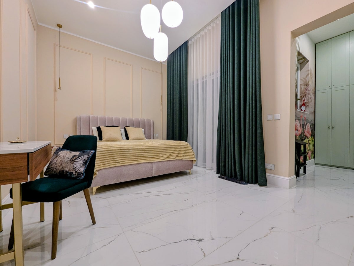A spacious bedroom is highlighted by a plush bed draped in soft, neutral tones. Elegant curtains frame large windows, allowing natural light to enter. A stylish desk and chair are positioned nearby, complementing the modern decor. A wardrobe is visible through an adjoining doorway.