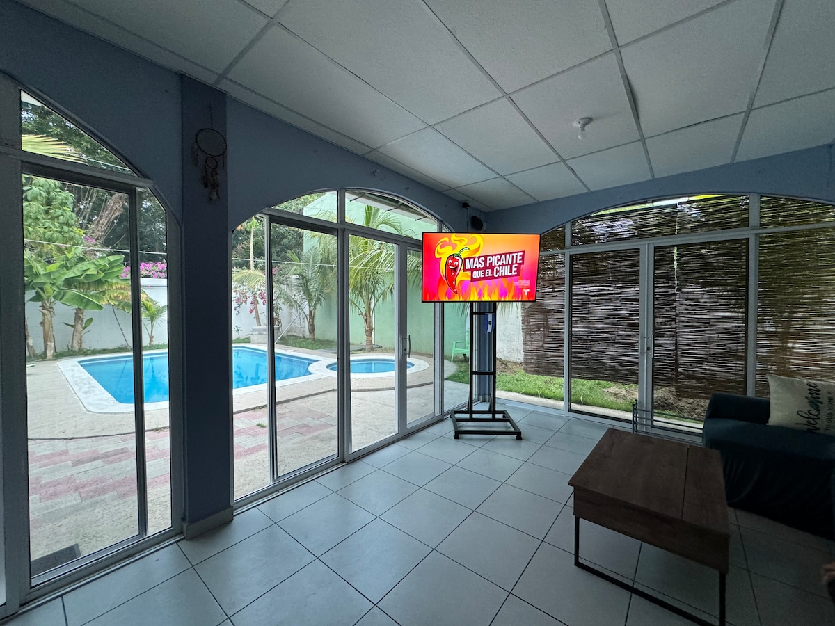 A large windowed space features a comfortable seating area with a view of the private pool outside. A flat-screen television is positioned on a stand, displaying colorful content. Natural light filters through the glass doors, illuminating the room's tiled flooring.