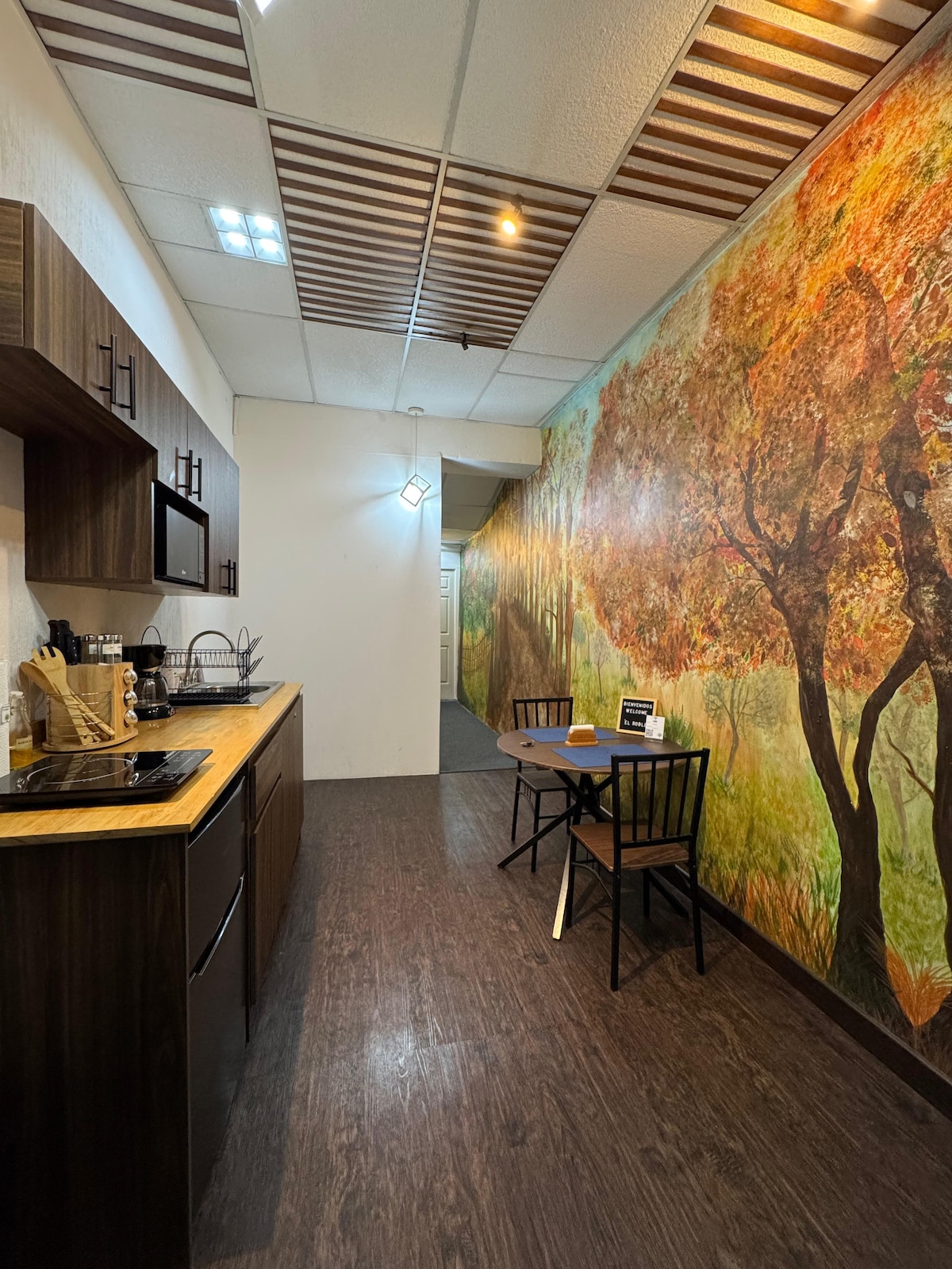 A functional kitchen area is equipped with a microwave, coffee maker, and various cooking utensils. A small table with two chairs is situated beside a vibrant mural featuring autumn trees, enhancing the ambiance of the space. Ambient lighting fixtures add to the overall atmosphere.