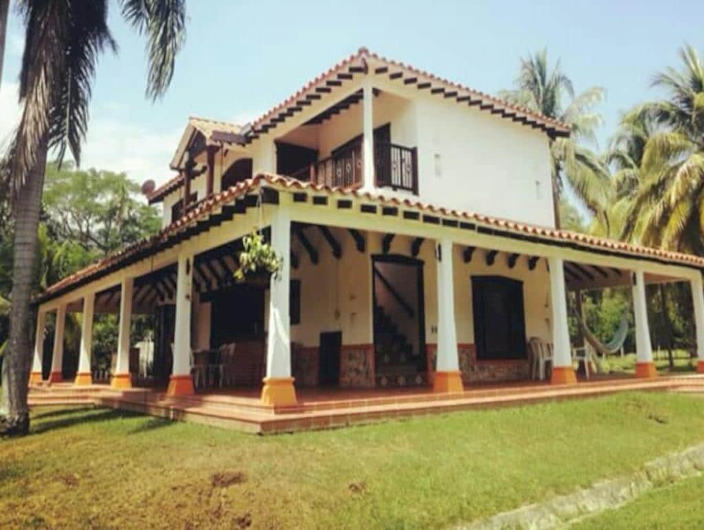 A spacious two-story finca is set amidst lush greenery, featuring a tiled roof and a wide veranda. Several outdoor chairs are positioned around the porch, and a staircase leads to the upper level. Palm trees and manicured lawn enhance the tropical ambiance.