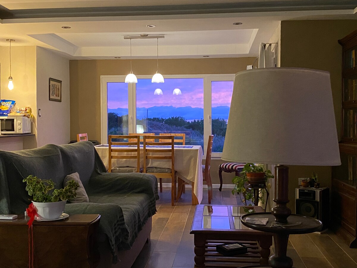 A living area features a comfortable sofa and a wooden dining table set against large windows showcasing a sunset view. A floor lamp is positioned nearby, providing warm lighting, while greenery adds a touch of nature to the space.