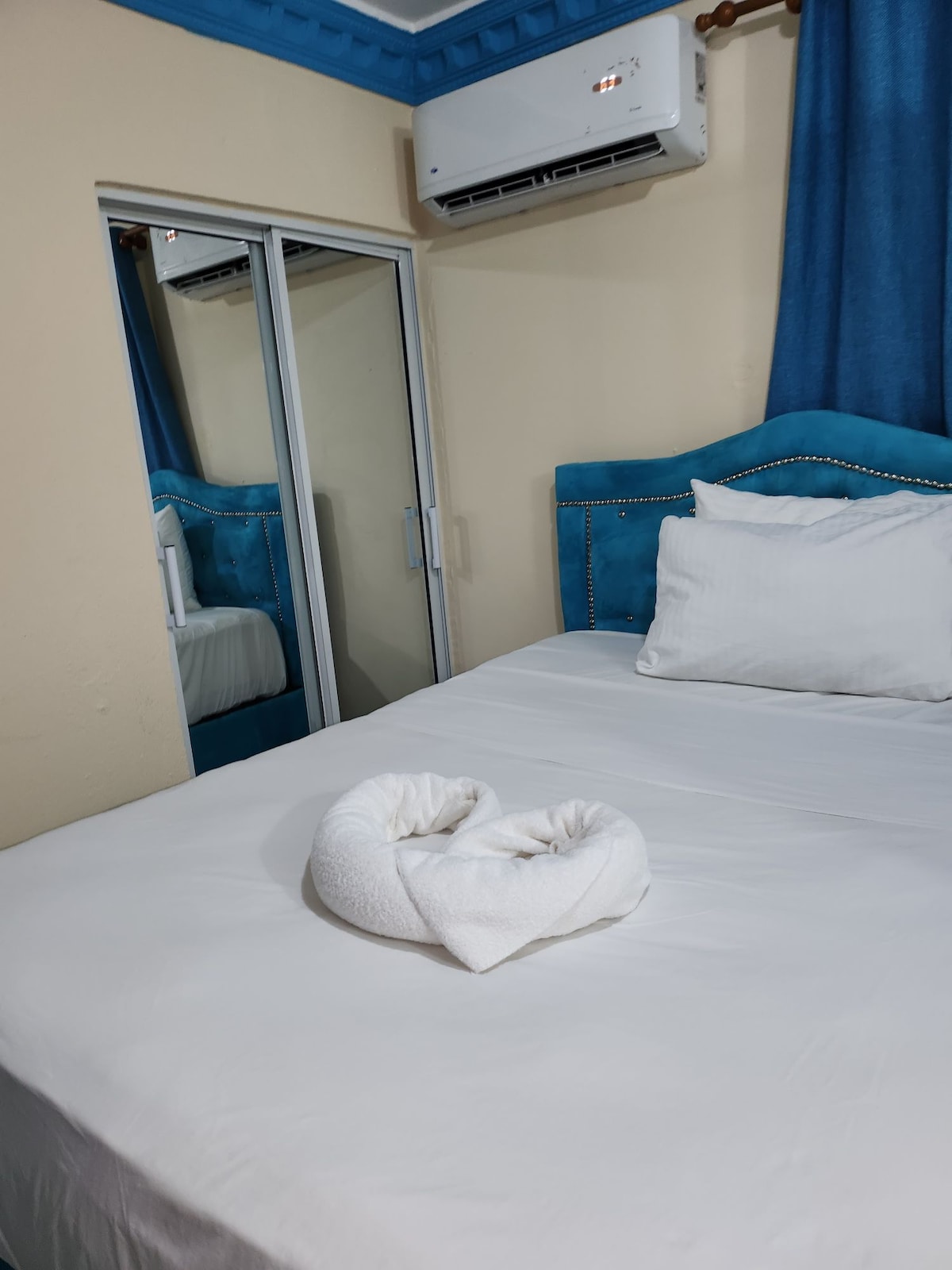 A spacious bed is adorned with white linens and a neatly arranged towel in a heart shape. A mirrored closet reflects the calming blue headboard and light blue curtains, creating a sense of openness in the room.