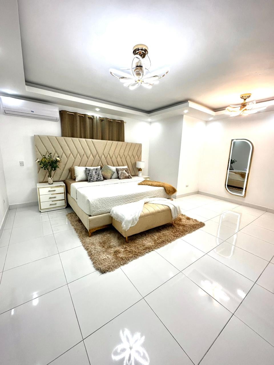 A spacious bedroom is presented, featuring a neatly made bed against a textured headboard. Soft lighting is provided by decorative ceiling fixtures. A large mirror stands against the wall, and a cozy area rug is positioned underfoot, contributing to a clean and modern aesthetic.