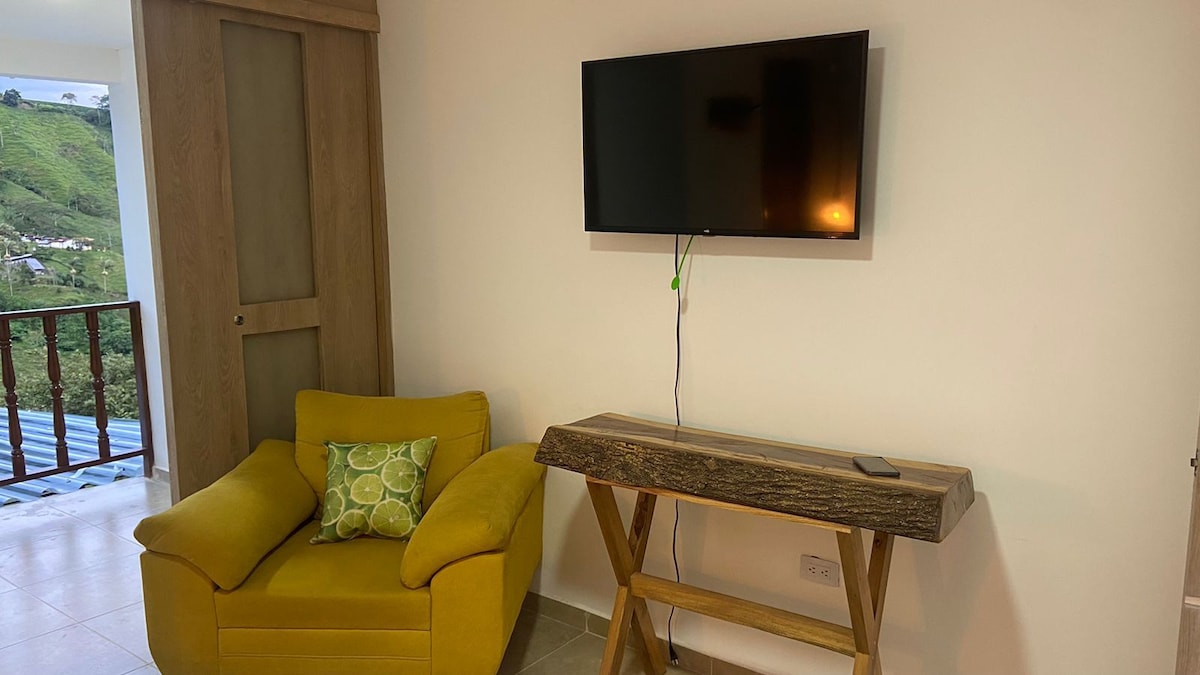 A cozy corner is highlighted by a yellow armchair featuring a decorative green pillow. A wooden console table stands nearby, supporting a flat-screen television mounted on the wall. Natural light floods the room, enhancing the neutral color scheme.
