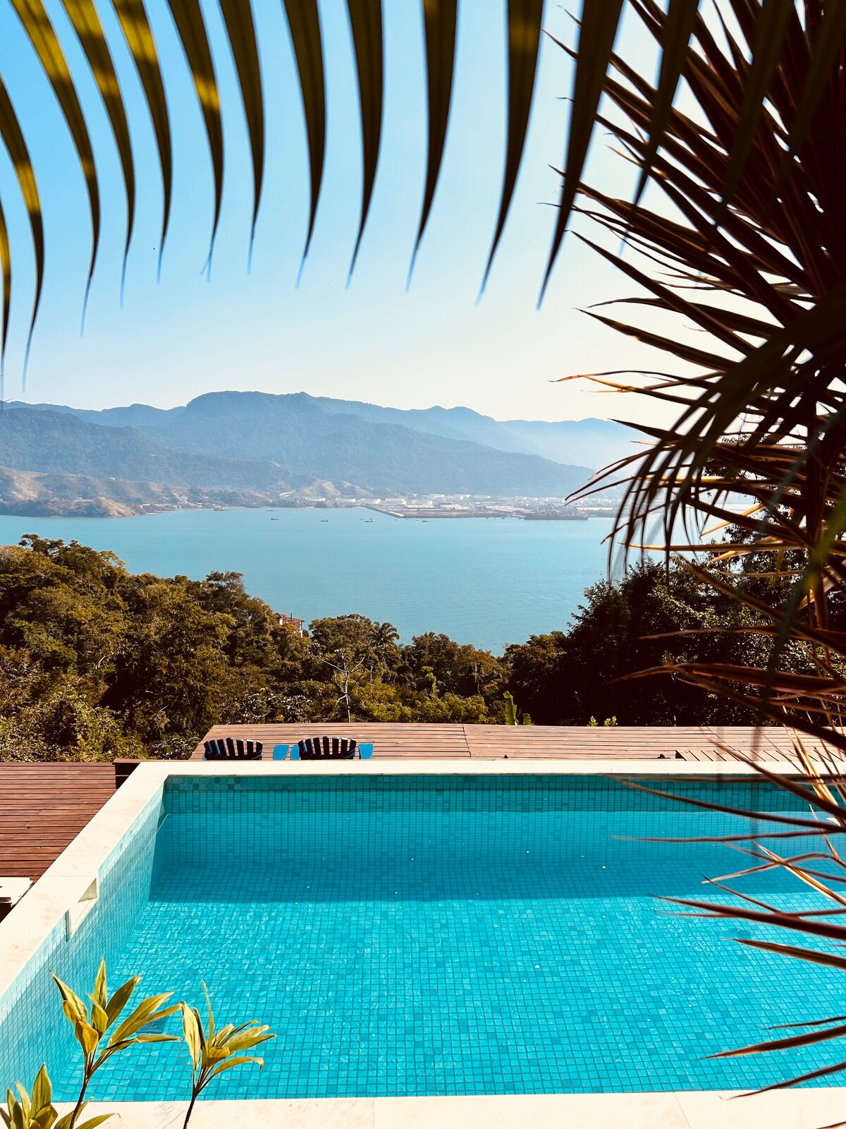 The image captures a sparkling pool framed by lush greenery, with the ocean and mountains visible in the background. Two lounge chairs are positioned by the poolside, offering a serene view of the landscape. Sunlight reflects off the water, enhancing the tranquil atmosphere.