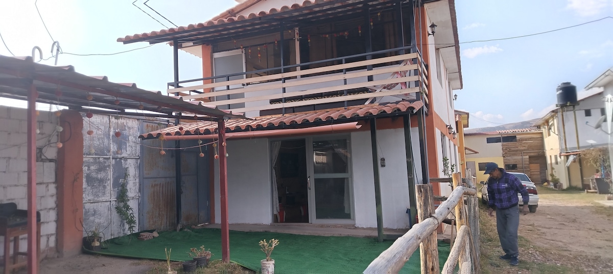 A two-story home is presented, featuring a spacious porch with a railing. Large glass sliding doors provide access to the interior. The front yard has a green lawn, bordered by a wooden fence, and is complemented by a few ornamental plants.