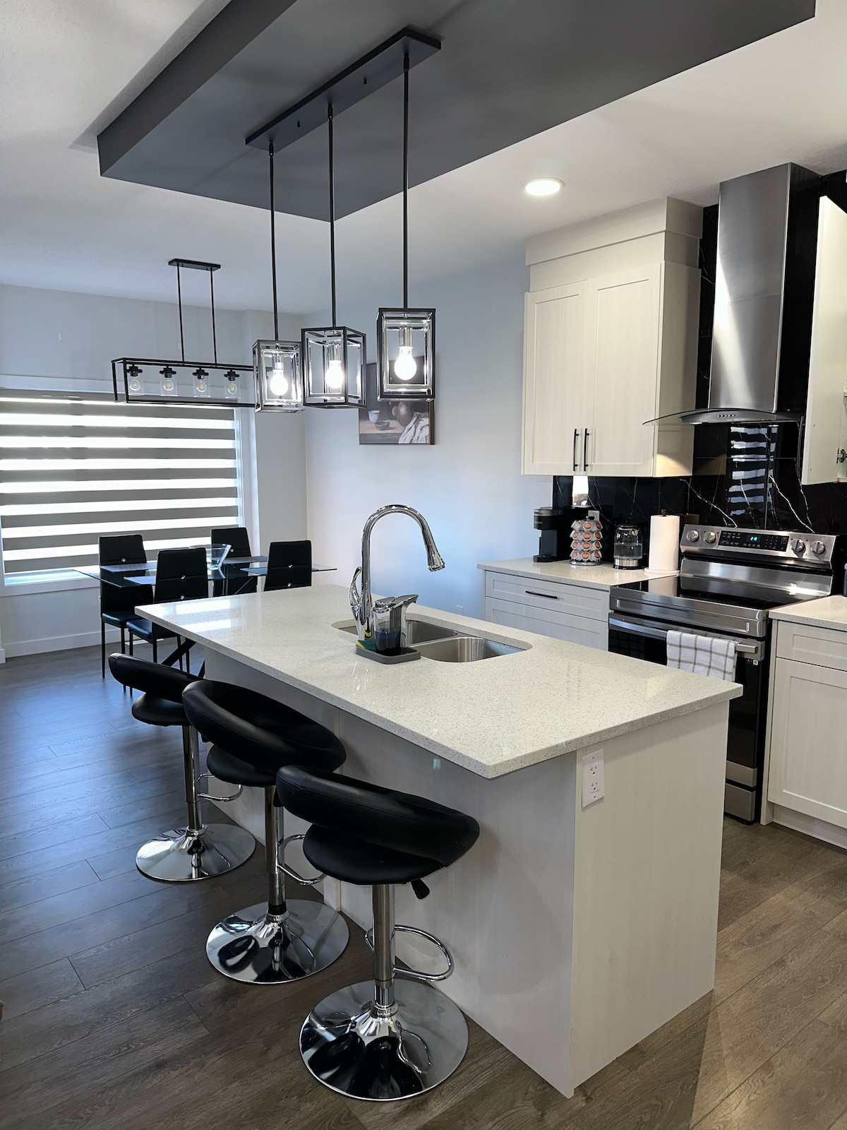 A modern kitchen space is highlighted by sleek cabinetry and stainless steel appliances. An island with a white countertop features three black bar stools. Overhead, three pendant lights provide illumination. A dining table with six chairs is situated nearby, complemented by a large window adorned with dual-tone shades.