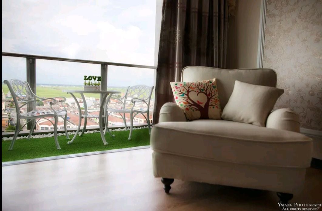 A cozy sitting area is featured, highlighted by a soft armchair and decorative cushions. A small balcony is visible, furnished with two chairs and a table, showcasing views of surrounding fields and distant buildings. Natural light fills the space, creating an inviting atmosphere.