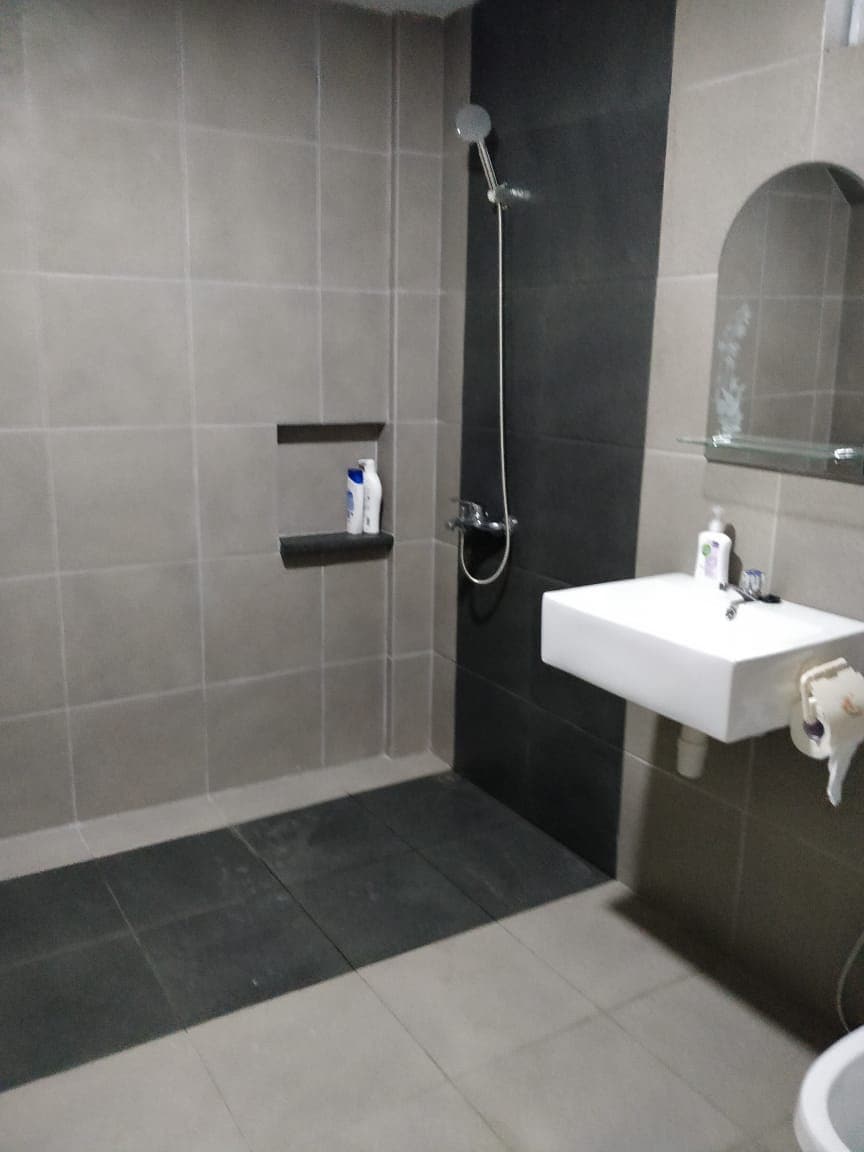 A modern bathroom features neutral-toned tiled walls and floors. A shower area is fitted with a handheld shower fixture, and a sleek white sink is mounted on a wall. A small shelf holds toiletries, while a mirrored shelf adds functionality.