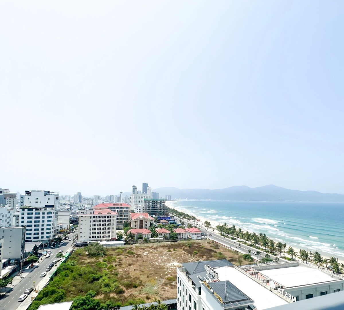 A panoramic view reveals the coastline along Mỹ Khê Beach, with a blend of modern buildings and greenery visible. The scene includes distant mountains and the expansive sea, allowing for an observation of the vibrant city landscape and the beachfront.
